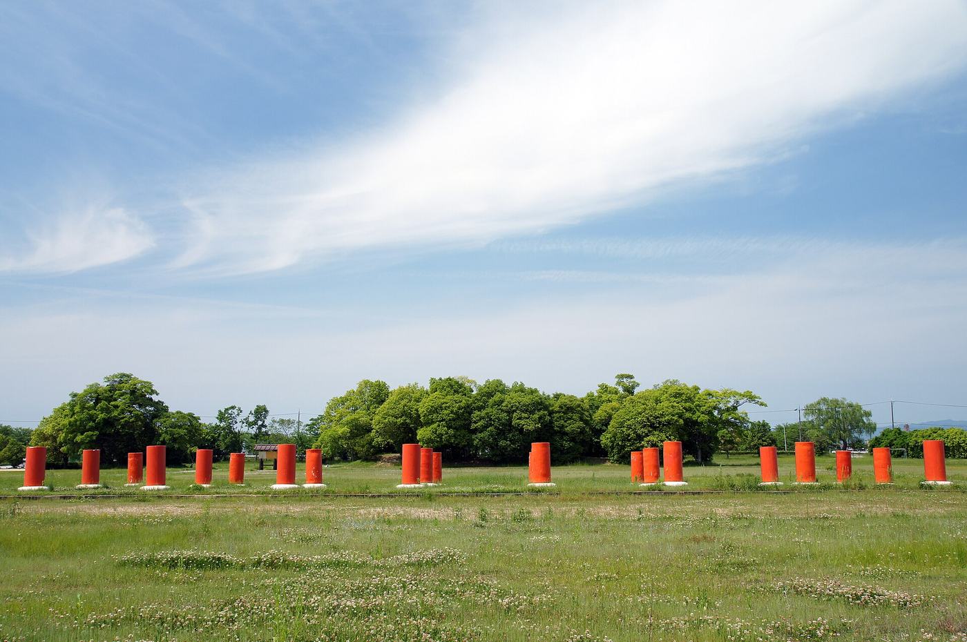 藤原宮跡:一整排橘紅色的圓柱矮樁立在白色方形基座上、橫向排開穿過大片草地,柱子最高只到成人腰部,柱列後方是一排樹叢與電線桿,再往後是低矮的遠山,天空佔了畫面上半部