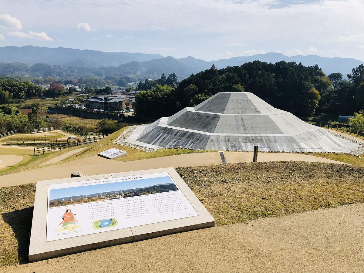 復元後的牽牛子塚古墳:淺灰白色切石一層層貼滿整座階梯狀墳丘、上緣收成平頂,四周鋪碎石與沙土步道並圍著木柵,前景地上斜放一塊寫著「国史跡 牽牛子塚古墳・越塚御門古墳」的解說板,背景是聚落、農地與層疊的遠山