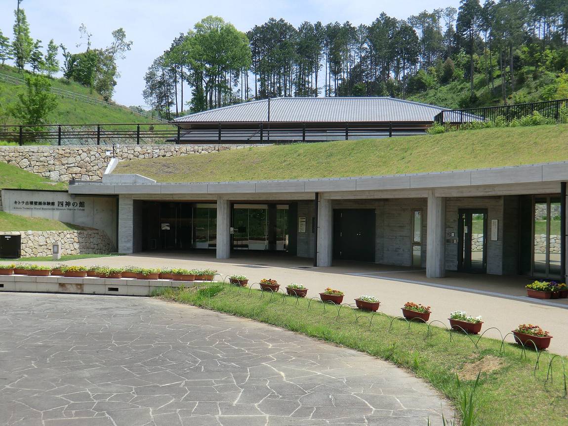 キトラ古墳壁画体験館「四神の館」的外觀:一棟嵌進草坡裡的清水混凝土建築,屋頂與後方山坡連成一片草地,左側牆面刻著日文與英文館名,前方是石板鋪面的圓弧步道與一整排花盆,建築後方的山坡上另有一棟灰瓦屋頂的小型建物