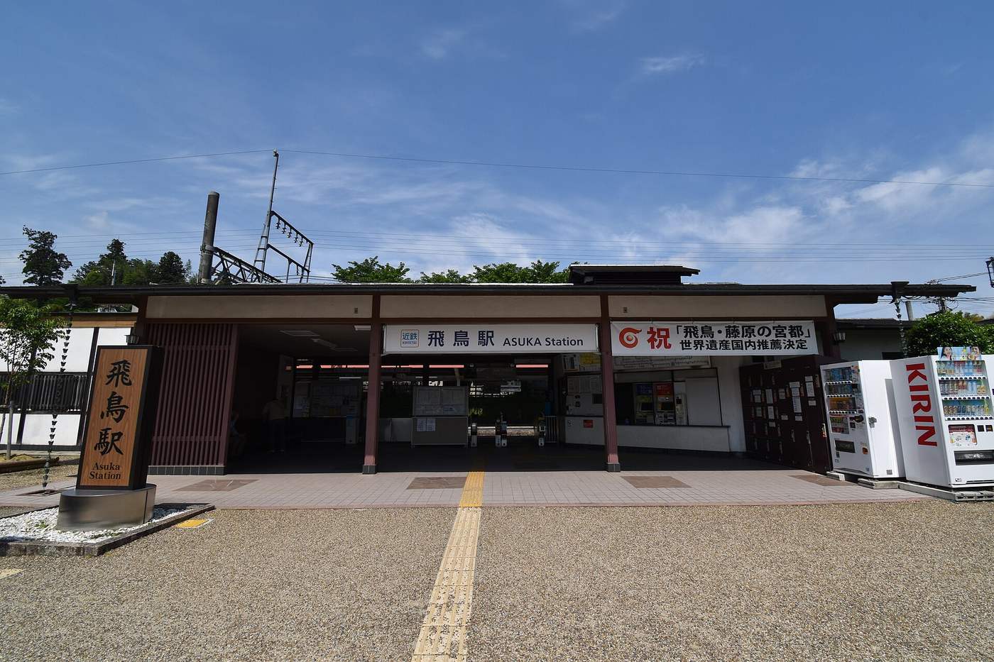 近鐵飛鳥駅站前:一棟低矮的和風站房,深色木柱與瓦簷下掛著白底招牌「近鉄 飛鳥駅 ASUKA Station」,右邊另掛一面寫著「祝『飛鳥・藤原の宮都』世界遺産国内推薦決定」的白色橫幅,站房內看得到售票機與剪票口,最右側並排兩台飲料販賣機,前方是碎石鋪面與黃色導盲磚