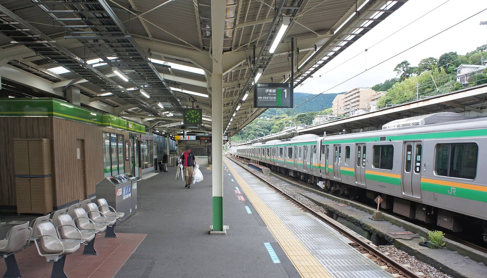 熱海站在來線月台,頭上懸掛的電子看板寫著「伊東線 1」與「伊東・伊豆急下田方面」,右邊軌道上停著一列綠色與橘色橫帶的 JR 電車,左邊是綠色木紋外牆的車站小賣店與一排灰色候車座椅,遠處是山坡與公寓大樓