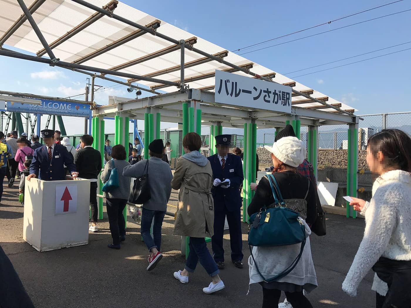バルーンさが駅出入口:白色鋼管與塑膠浪板搭成的臨時遮棚下掛著寫有「バルーンさが駅」的白底黑字站名牌,一整排螢光綠的鋼管柵欄圍出動線,兩名穿深藍色制服戴大盤帽的 JR 站員站在通道上人工驗票,左方藍色橫幅寫著 WELCOME,遊客成群穿過