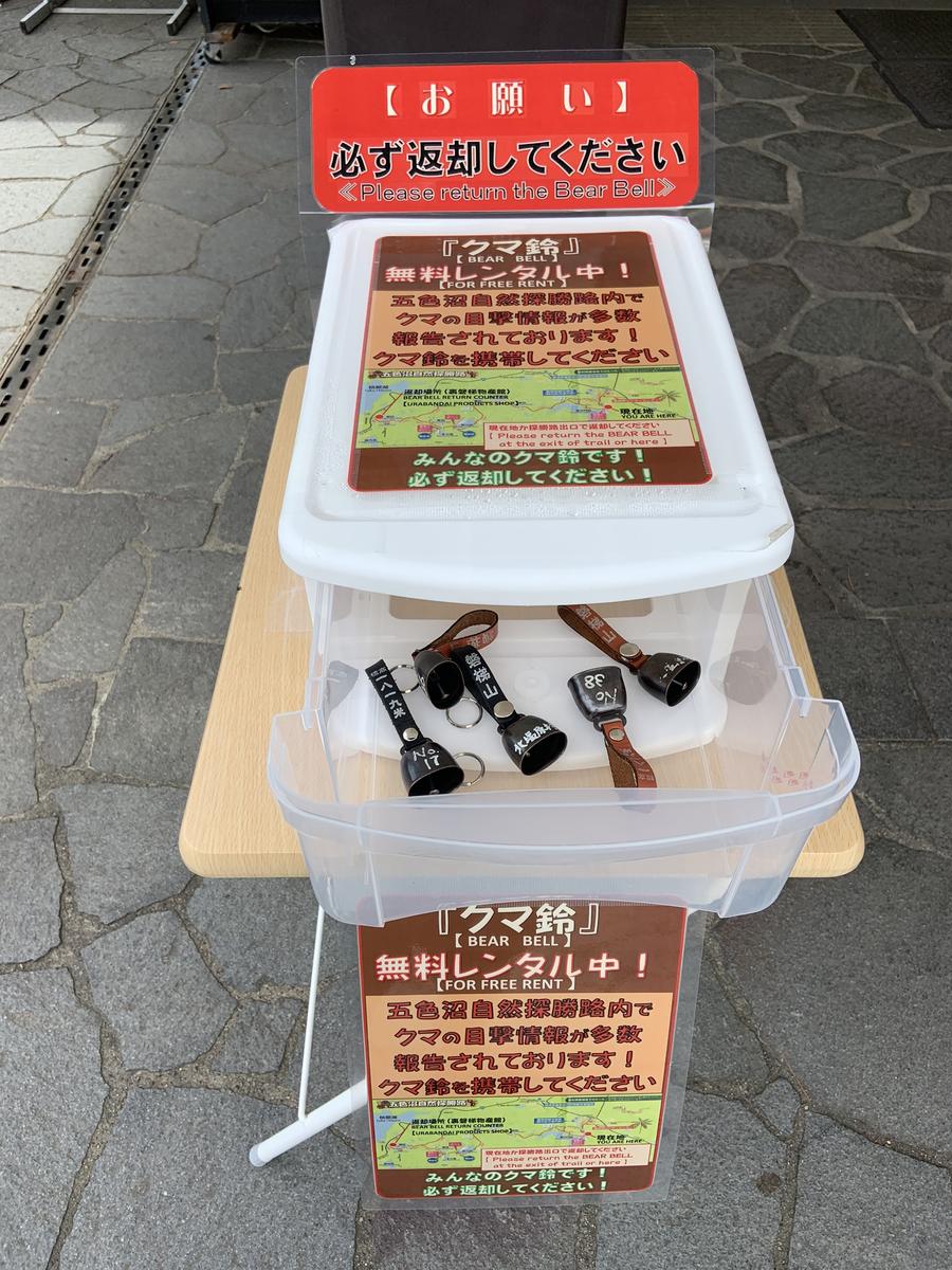 步道入口鋪石地面上的熊鈴免費出借台，木桌上放著透明抽屜箱，抽屜裡躺著四只繫皮帶、寫有編號的黑色鈴鐺，箱上與箱前各貼一張「クマ鈴 無料レンタル中！」海報，最上方紅底立牌寫著「必ず返却してください／Please return the Bear Bell」