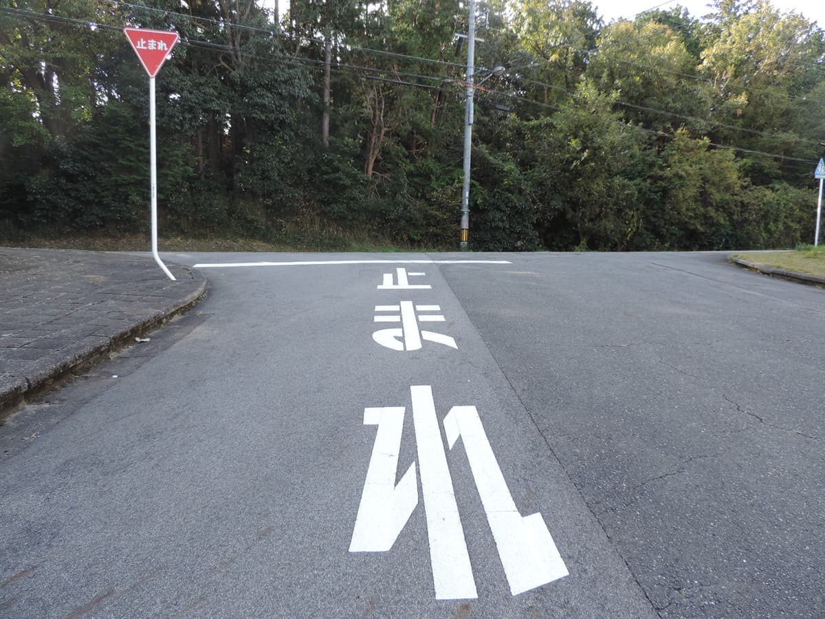 日本住宅區小路口的一時停止:路面上用白漆漆著「止」「ま」「れ」三個大字,字的前方橫著一道白色停止線,左邊人行道邊立著一支紅色倒三角形的「止まれ」標誌牌,路口對面是一整片樹林