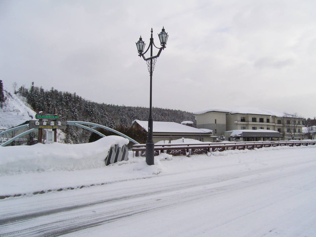 三月初的白金溫泉入口:整條車道被壓實的雪蓋住,橋邊雪堆比欄杆還高,左邊有寫著「白金温泉 SHIROGANE SPA」的木牌和藍綠色拱橋,右後方是屋頂積雪的旅館