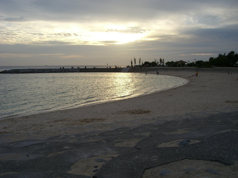 傍晚的北谷 Sunset Beach,弧形沙灘圍著平靜的海灣,雲層後的太陽在海面映出一道光,遠處防波堤上坐著幾個小小的人影
