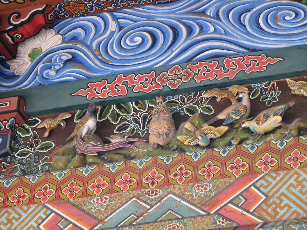 秩父神社社殿的彩色木雕近景，中央是一隻身體朝前、頭轉向正面的貓頭鷹站在綠色苔石上，左右各有數隻彩繪小鳥與長尾鳥；上方是藍白相間的波浪雕刻，下方是金底六角龜甲紋與花草彩繪