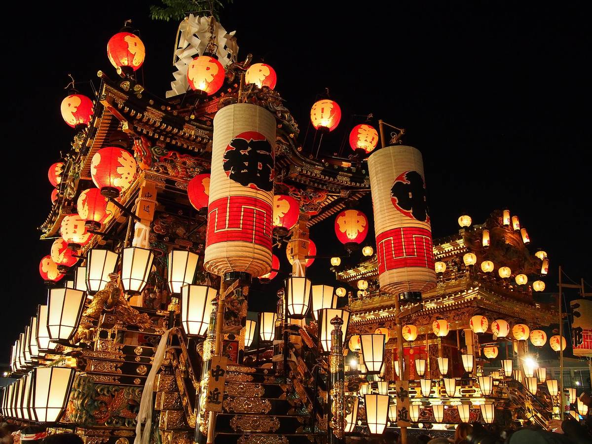 兩座掛滿提灯的秩父夜祭笠鉾在夜裡並排，前方那座掛著寫「中近」的木牌，車頂旁立著兩支白底紅帶的大型圓筒提灯，下層排滿方形白提灯，車體是彩色木雕加金箔，背景是全黑的夜空