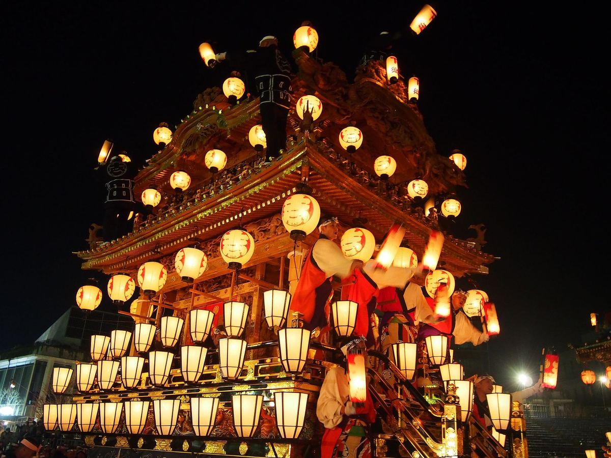 秩父夜祭的屋台在夜裡由下往上仰拍，兩層屋簷與整面欄杆掛滿數十盞亮著的紅色圓提灯與白色方提灯，屋頂上站著兩名穿黑色半纏的男子，欄杆邊幾個穿紅白裝束的人正把手提提灯往外揮