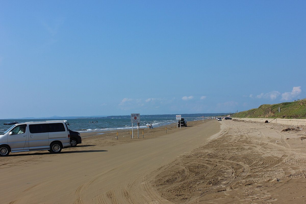 千里浜渚公路,一片被海浪壓實的寬闊沙灘直接當車道用,沙面上滿是輪胎壓出的痕跡,左側停著一輛廂型車,遠方沙灘上有幾輛車與零星釣客,右側是長草的沙丘,天空湛藍