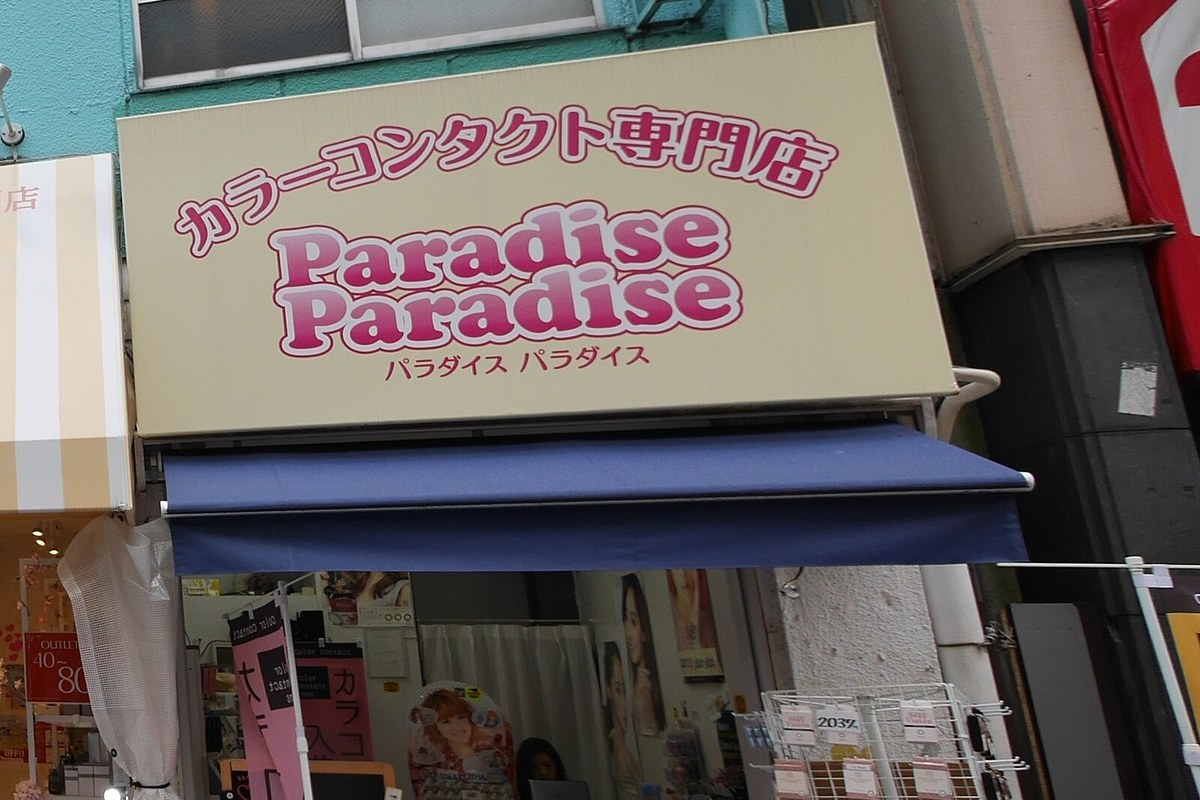 東京下北澤街邊一間彩色隱形眼鏡專門店,店招寫著「カラーコンタクト専門店」,遮陽棚下是店頭的商品展示架
