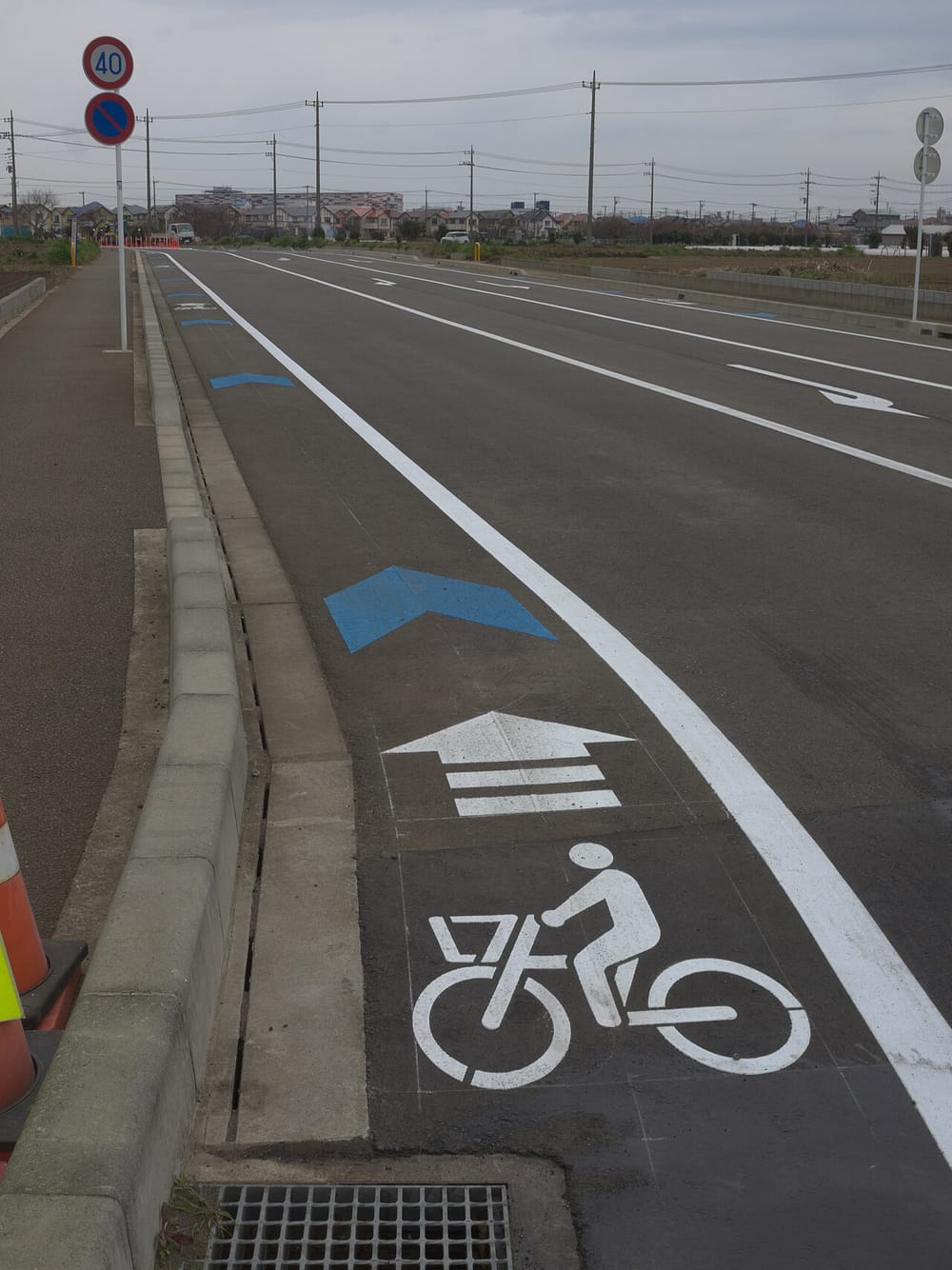 日本道路左緣的自転車ナビマーク:白色單車圖示與三道人字紋的前進箭頭畫在路肩白線內側,前方接一枚藍色人字形箭頭,右側對向車道的白色箭頭指向相反方向