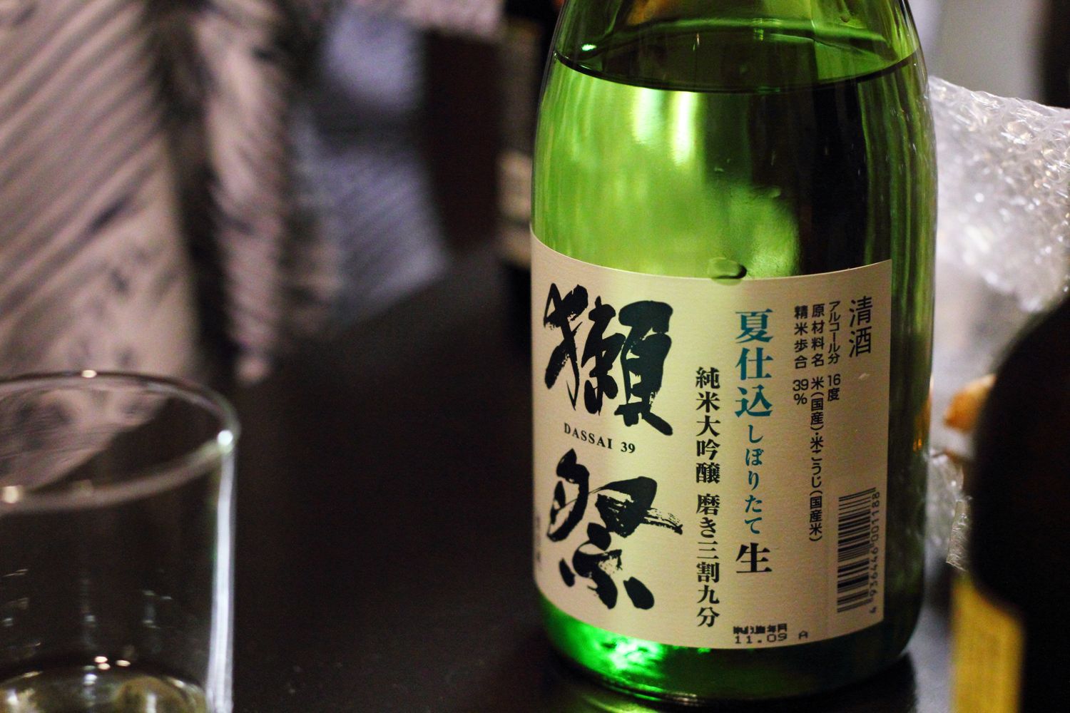 一支綠色玻璃的獺祭酒瓶擺在深色桌面上，瓶身貼著米白色直式標籤，右起以毛筆體寫著大大的「獺祭」二字，下方小字印著 DASSAI 39；標籤中段由右至左直排著「夏仕込 しぼりたて 生」與「純米大吟醸 磨き三割九分」，其中「生」字單獨一個字、字級明顯放大，再往右是清酒、原材料名、精米歩合 39%、アルコール分 16 度等成分欄與條碼。畫面左前方有一只空的玻璃杯，右側可以看到一疊氣泡布