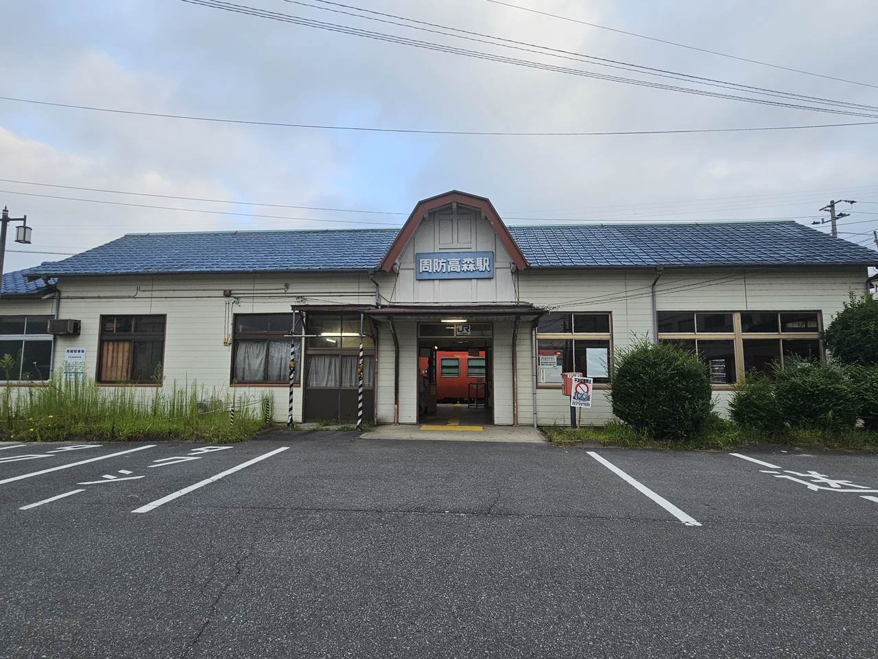 周防高森駅站體正面：一棟米白色木造平房，藍色瓦片屋頂，正中央的三角形山牆上掛著藍底白字的「周防高森駅」站名牌，入口門楣上是白色的 JR 標誌；穿過敞開的出入口可以看到月台上停著一輛朱紅色柴油列車。屋子兩側的窗前長著及腰的雜草，右側立著一塊紅白色的告示牌。建物前方是一片畫著白色停車格線的柏油空地，格子裡一輛車都沒有，也沒有任何排班車輛