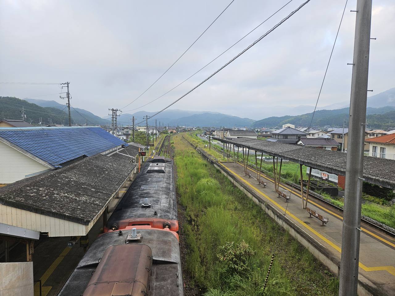 從跨線橋上俯瞰周防高森駅：畫面左下是兩節朱紅色柴油列車停在藍瓦站房旁的月台邊，右側是另一座老舊木柱雨棚的月台，上面排著幾張長椅、空無一人；兩座月台之間的軌道與空地長滿及膝的綠色雜草。再往遠處是低矮民宅組成的小鎮，一路延伸到被雲霧壓著的連綿山稜，天空是灰白色的陰天