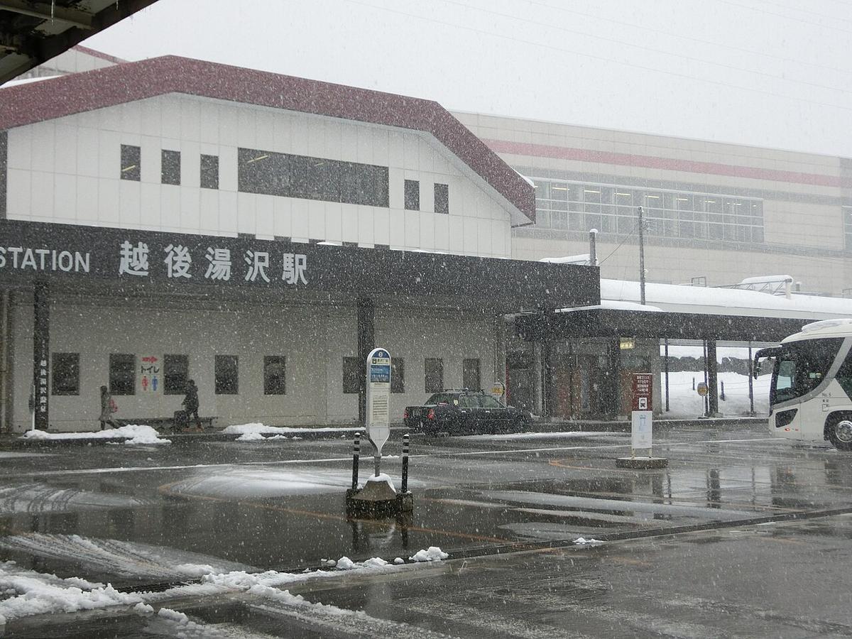 下雪中的越後湯沢站東口：紅色斜屋頂的白色低矮站房，黑色招牌寫著「越後湯沢駅」，後方是新幹線的高架站體；站前廣場濕滑、路邊有積雪，中間立著一根公車站牌，右側停著一輛白色大型巴士，左邊有兩個走過的小小人影