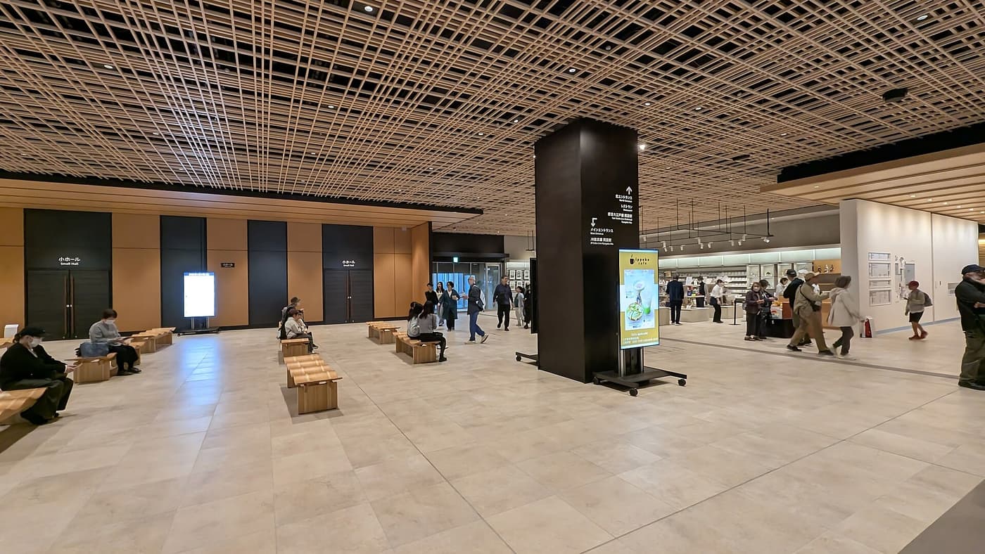 江戶東京博物館 1 樓大廳:木格柵天花板下是一片開闊的米色石材地板,散放著數張淺色木長凳並有人坐著,中央一根深色方柱印著往北エントランス、レストラン、都營大江戶線兩國站與 JR 總武線兩國站的指示箭頭,右側是明亮的商店區