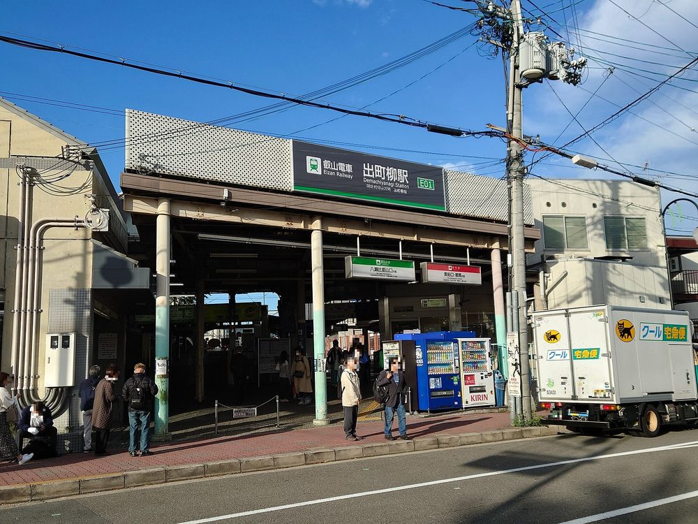 叡山電車出町柳站的地面站房:上方黑底招牌寫「叡山電車 出町柳駅 E01」,入口棚下掛兩塊方向牌,綠色是「八瀬比叡山口方面」、紅色是「貴船口・鞍馬方面」,路邊有人等候(臉部已由上傳者打馬賽克)