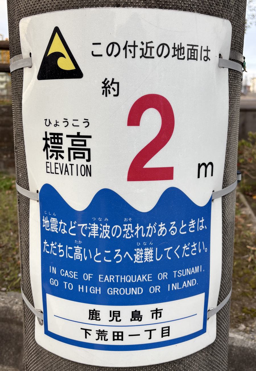 綁在電線桿上的白色標高表示板：上半是黃黑色的海嘯警告三角圖示與「この付近の地面は 約 2 m」，紅色大字的 2 特別醒目，左側標著「標高 ELEVATION」；下半藍底白字寫著「地震などで津波の恐れがあるときは、ただちに高いところへ避難してください。」並附英文 IN CASE OF EARTHQUAKE OR TSUNAMI, GO TO HIGH GROUND OR INLAND.，最下方標示鹿児島市 下荒田一丁目