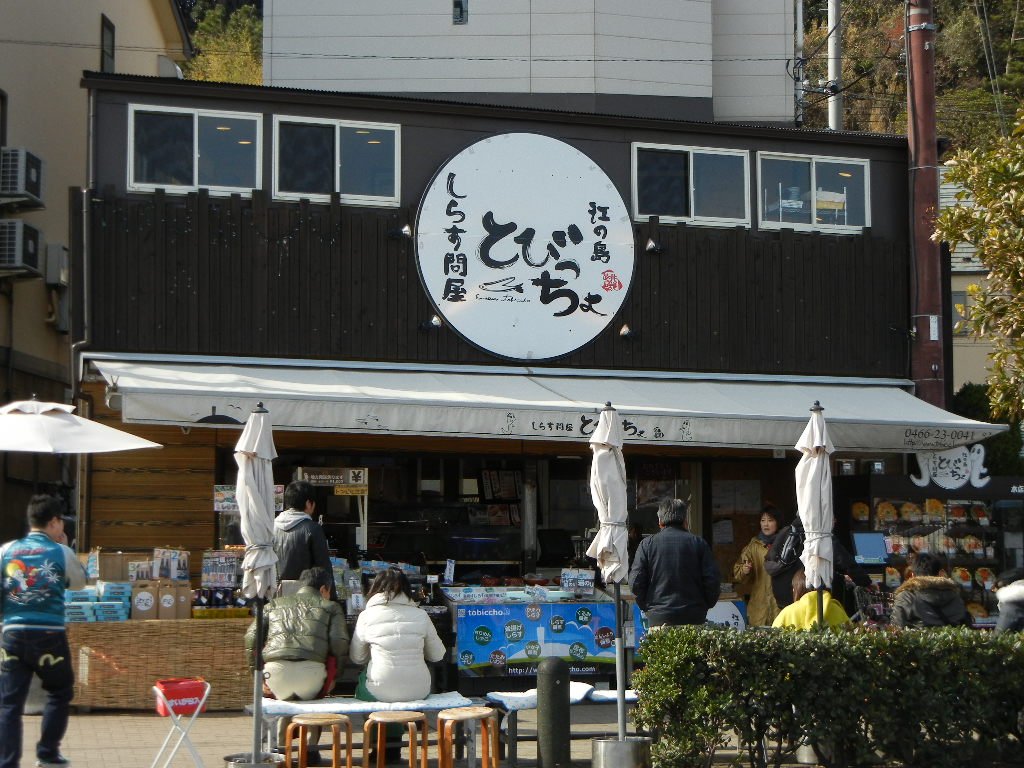 江之島とびっちょ本店的店面,黑色木造外牆掛著白色圓形「しらす問屋 とびっちょ」招牌