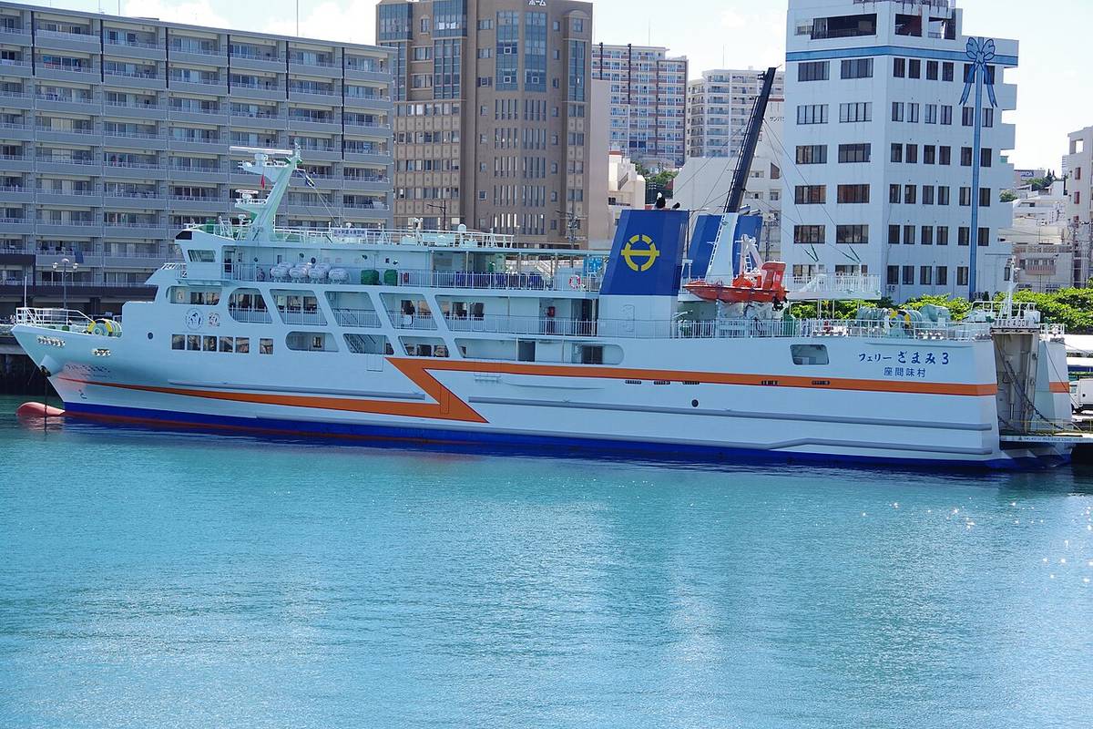 停靠在那霸泊港的渡輪「フェリーざまみ3」，白色船身有一道橘色與深藍色的折線塗裝，船側寫著座間味村，背景是那霸的公寓大樓