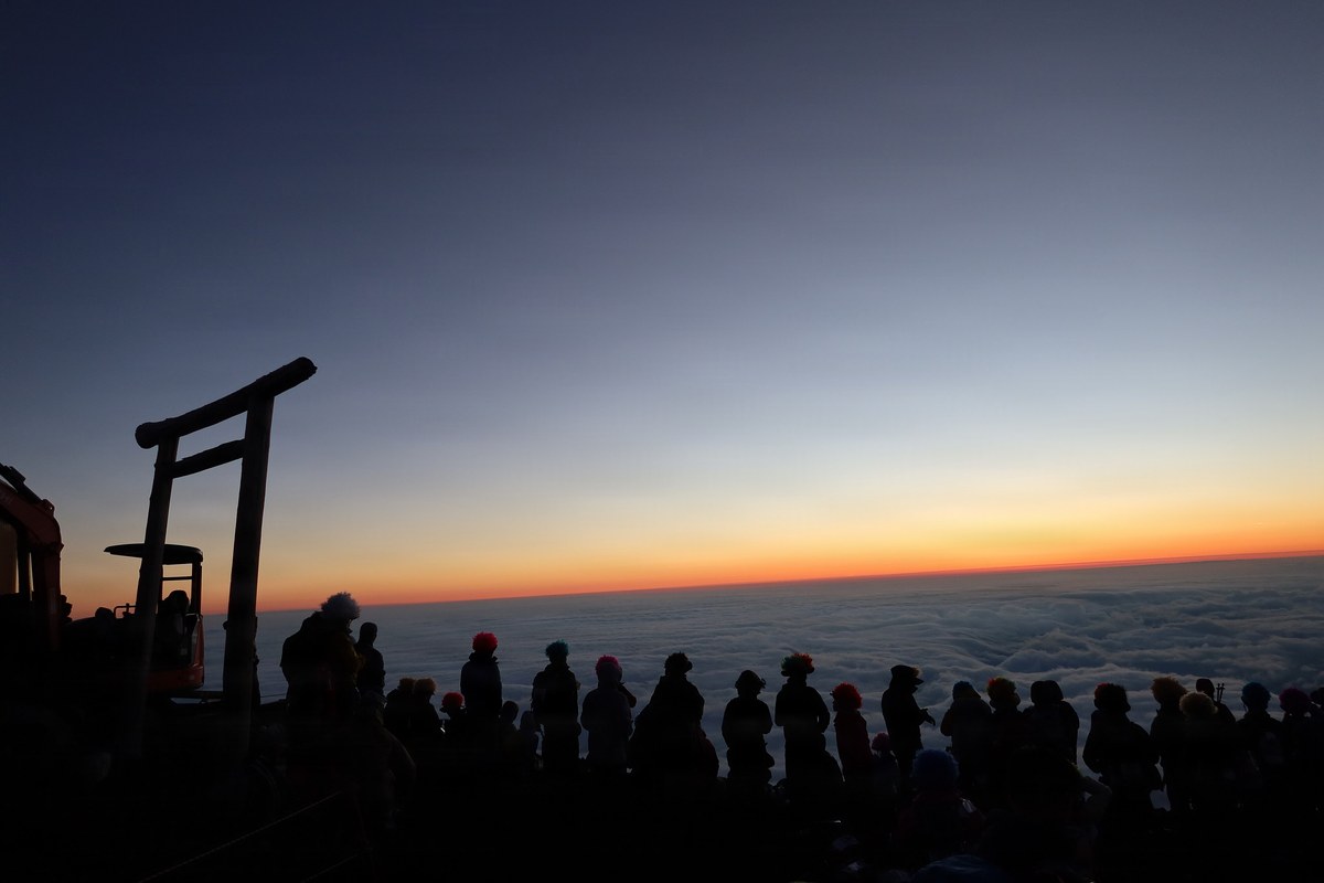 日出前的富士山頂，一長排登山者背對鏡頭沿著崖邊站成一列剪影，左側是木造鳥居的輪廓，前方雲海鋪到天邊
