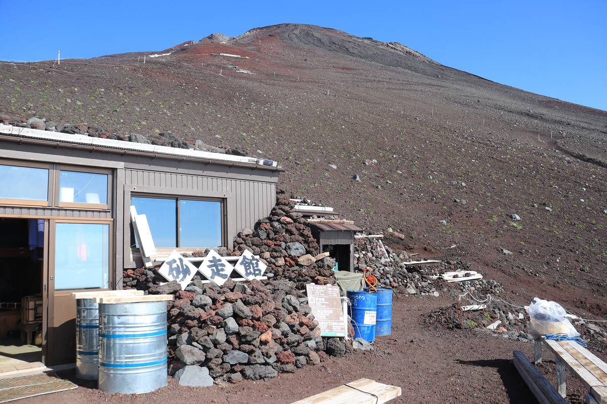 富士山山小屋「砂走館」的外觀，鐵皮屋身下方用黑紅色火山岩塊層層堆砌護牆，屋後是一整片沒有植被的火山礫斜坡