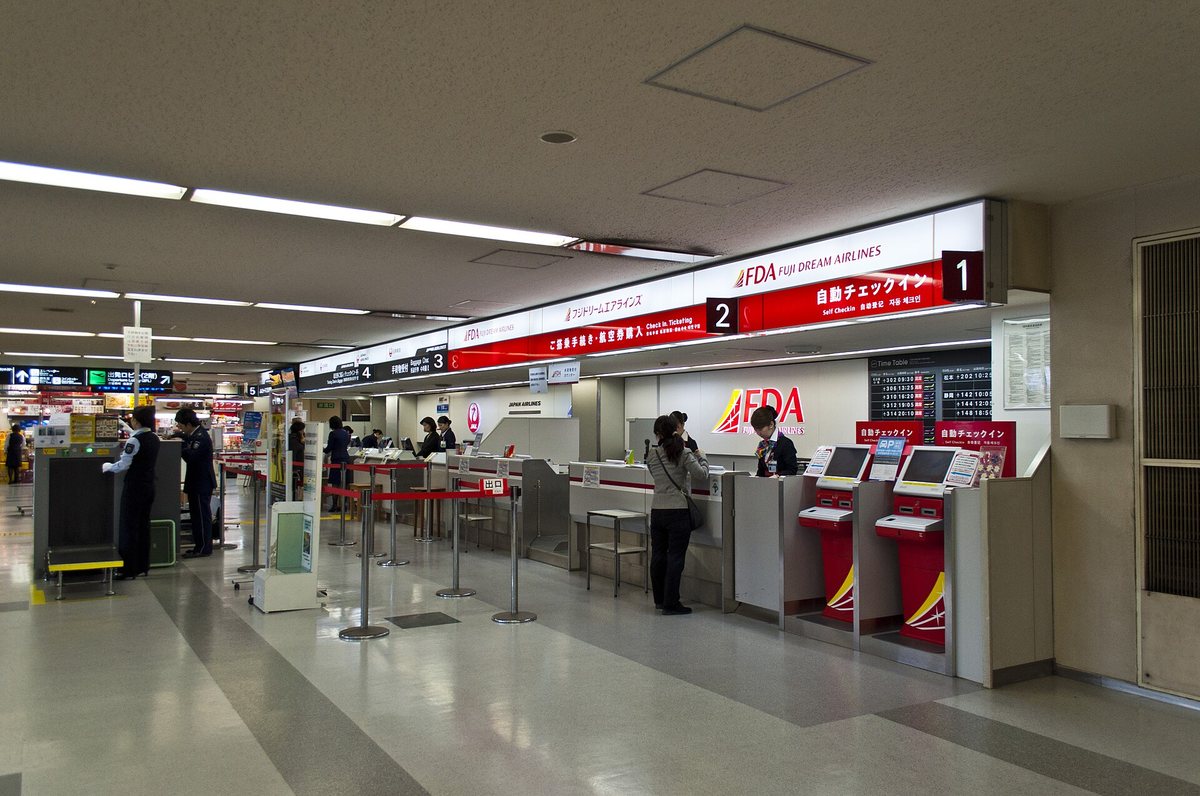 福岡機場國內線航廈的報到區,紅色橫式燈箱寫著「自動チェックイン Self Checkin 自動登記 자동 체크인」與編號 1、2,右側兩台紅色自助報到機,左半是日本航空的人工櫃檯與鶴丸標誌
