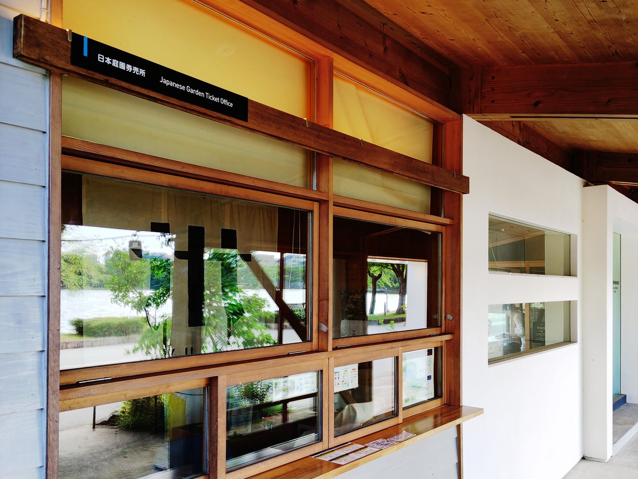大濠公園日本庭園的售票窗口:木造屋簷下一排木框玻璃窗,上方黑底白字的牌子寫著「日本庭園券売所 Japanese Garden Ticket Office」,窗台上攤著幾張摺頁,玻璃上貼著園內地圖與電子支付標誌
