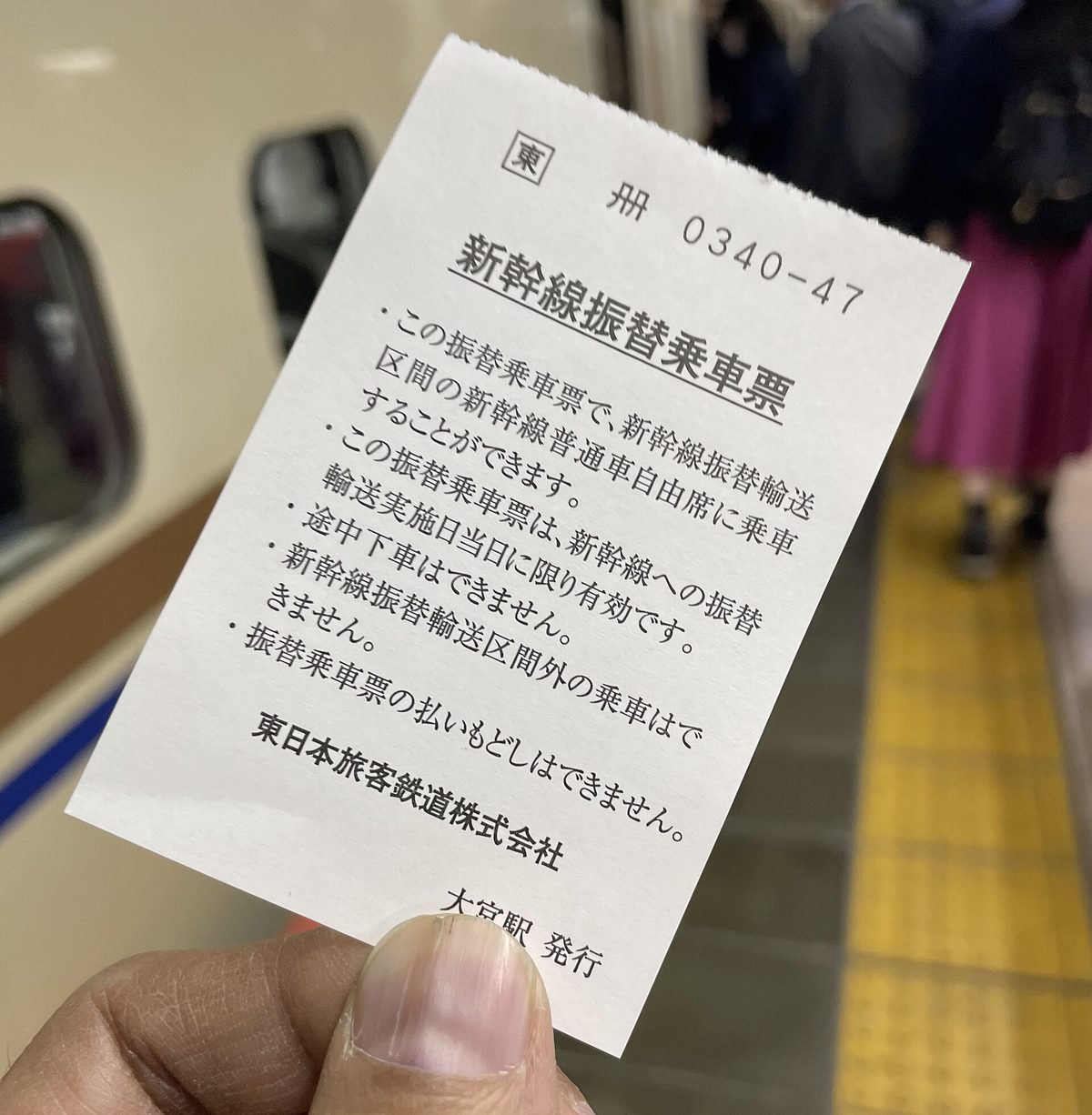手持一張 JR 東日本大宮站發行的新幹線振替乗車票，票面條列僅限實施日當日有效、不可途中下車、區間外不可乘車、不辦退款等條件，背景是月台上的新幹線列車與候車旅客