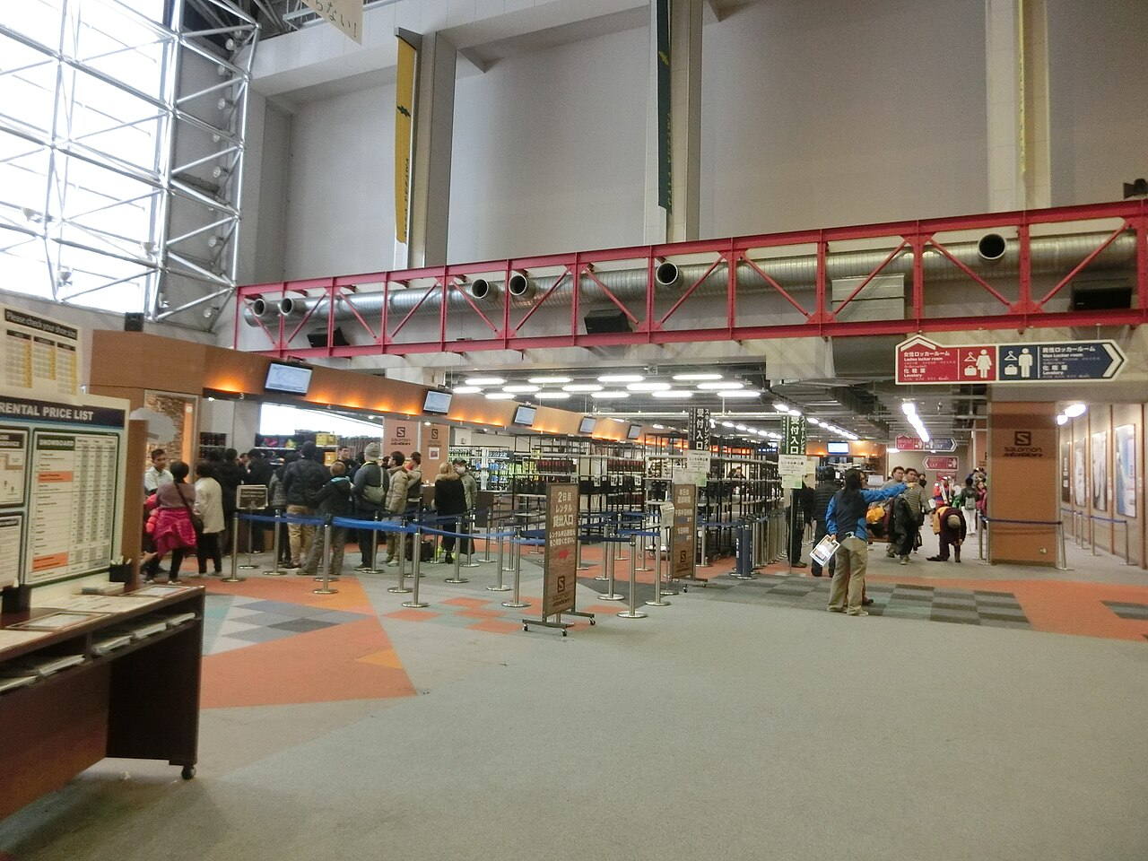 GALA湯沢滑雪場室內租借櫃檯，左側立著印有 SNOWBOARD 與 SKI 兩欄的 RENTAL PRICE LIST 價目看板，旅客沿藍色伸縮帶排隊等候受付