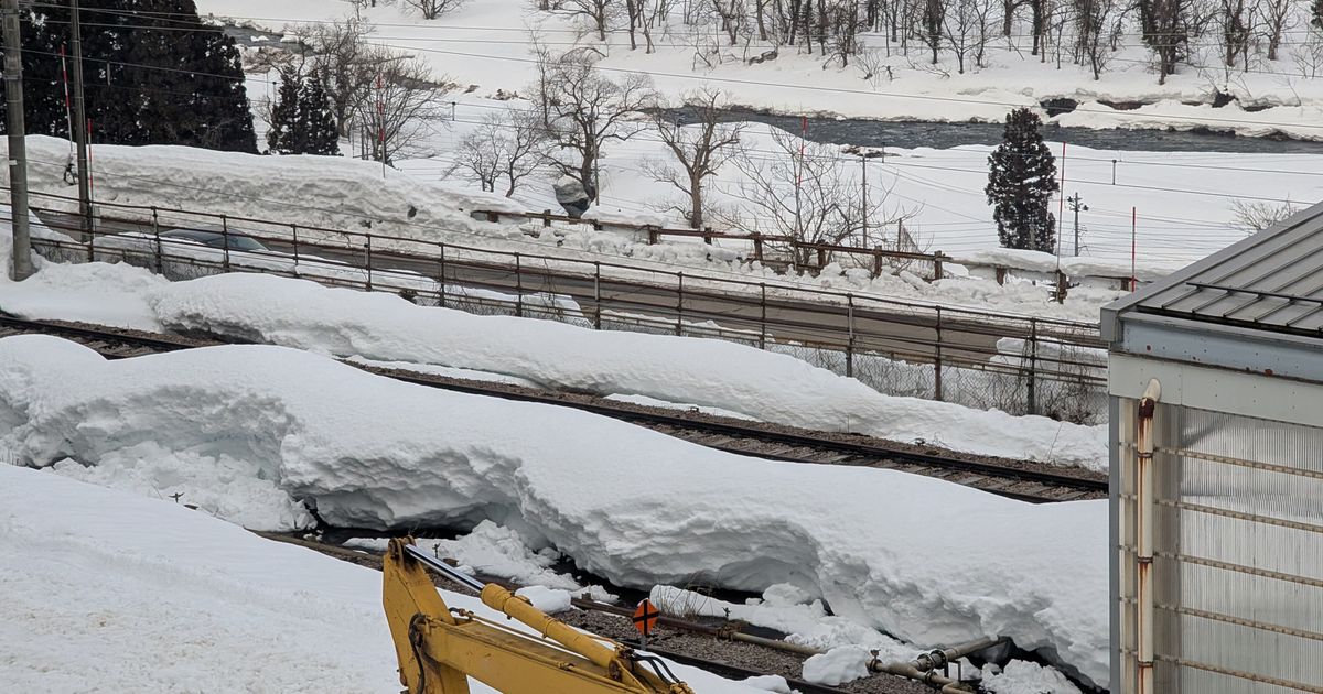 GALA湯沢駅旁的積雪鐵道：軌道間的雪堆成長條雪堤，護欄外是河流與覆雪樹林，前景有一截黃色挖土機手臂