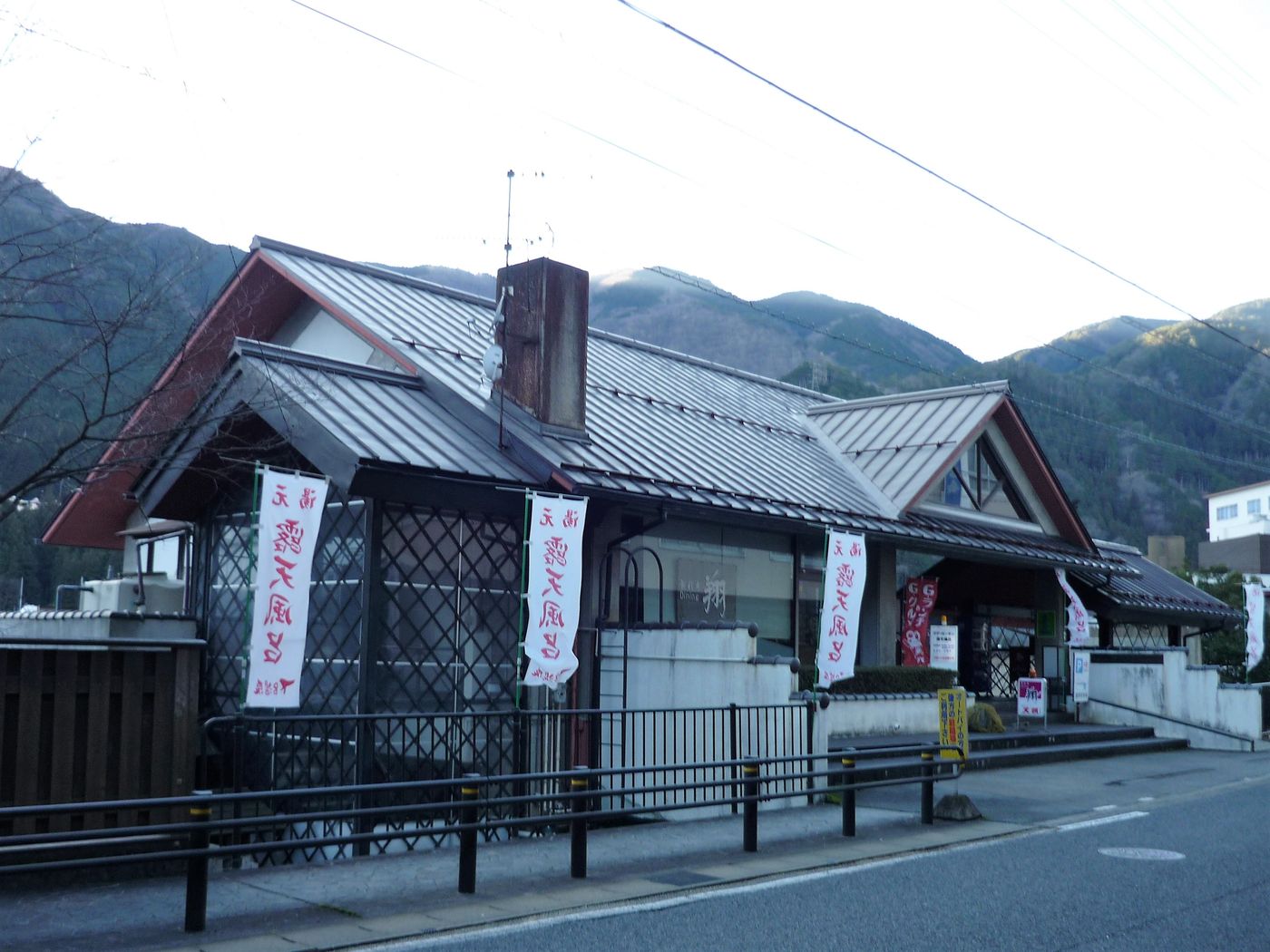 クアガーデン露天風呂的入口：瓦頂的和風平房，門前立著幾面寫著「露天風呂」的白色旗幟，背後是山
