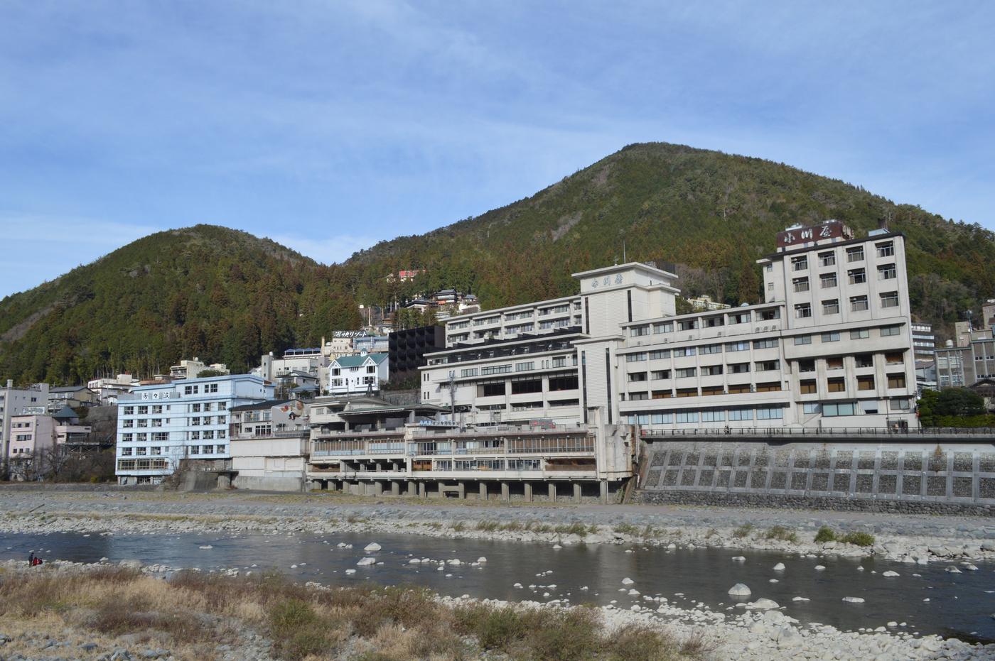 飛驒川河灘對岸，沿著河岸蓋起的多層溫泉旅館，最高那棟屋頂招牌寫著「小川屋」，背後是兩座長滿樹的山
