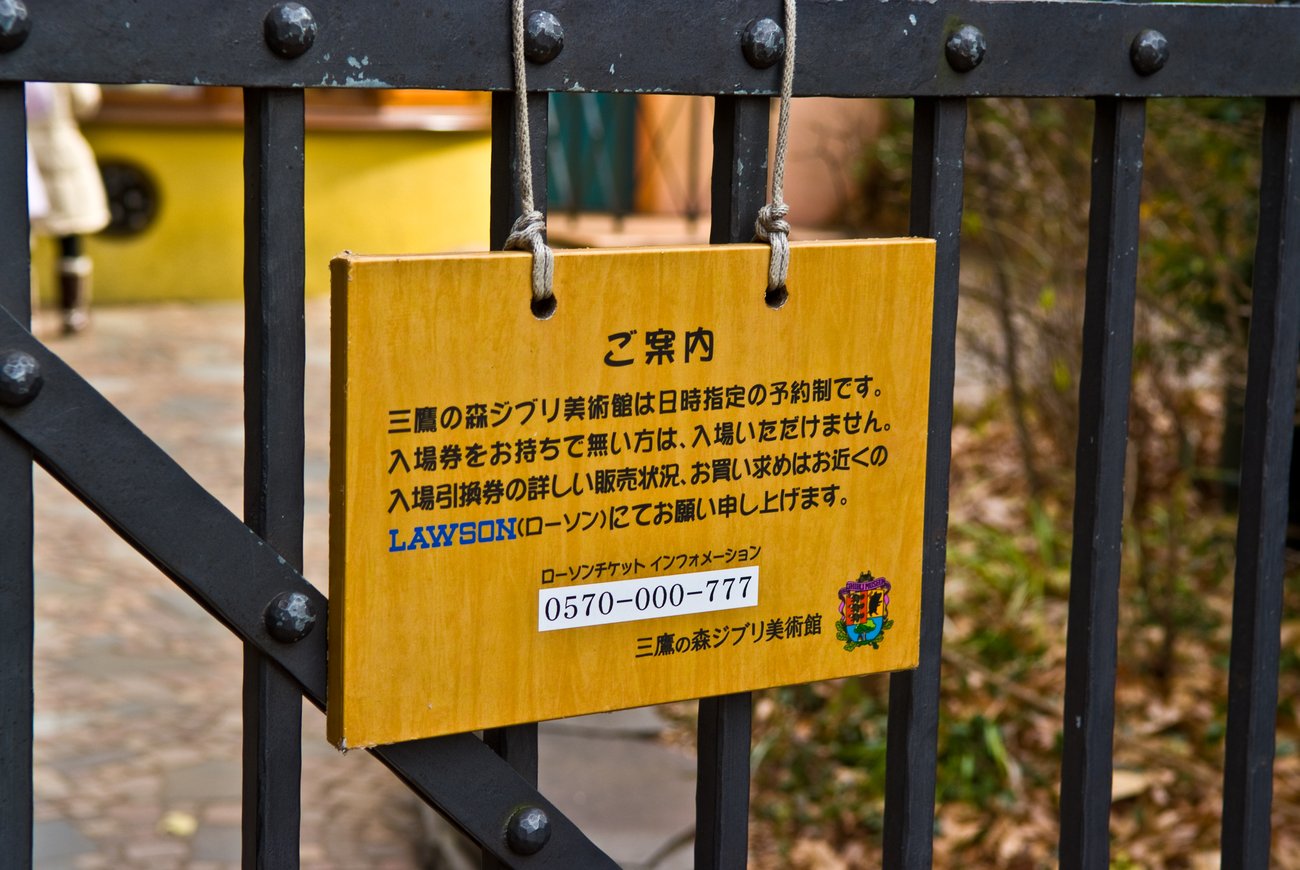 黑色鑄鐵柵門上用兩條麻繩掛著一塊黃褐色木牌，牌面黑字直式標題「ご案内」，內文四行日文寫著美術館採日時指定預約制、沒有入場券的人無法入場、詳細販售狀況與購買請洽附近的 LAWSON；LAWSON 幾個字以藍色印刷，下方框著電話號碼 0570-000-777，右下角是美術館的彩色徽章與「三鷹の森ジブリ美術館」落款；柵門後方背景是模糊的黃色牆面與石板地