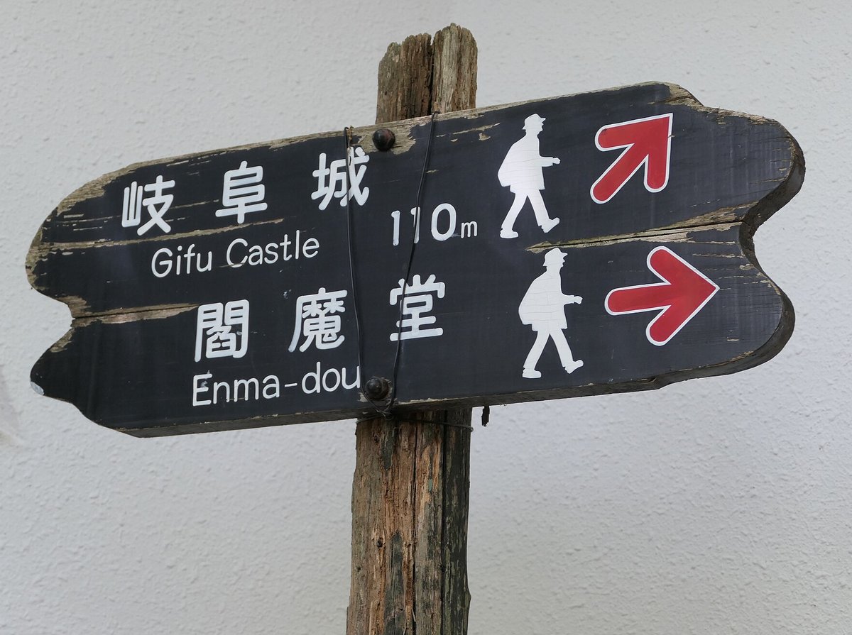 金華山山頂的黑色木製路標:上排寫「岐阜城 Gifu Castle 110m」配右上紅色箭頭,下排寫「閻魔堂 Enma-dou」配向右紅色箭頭,兩排都畫著白色步行人形