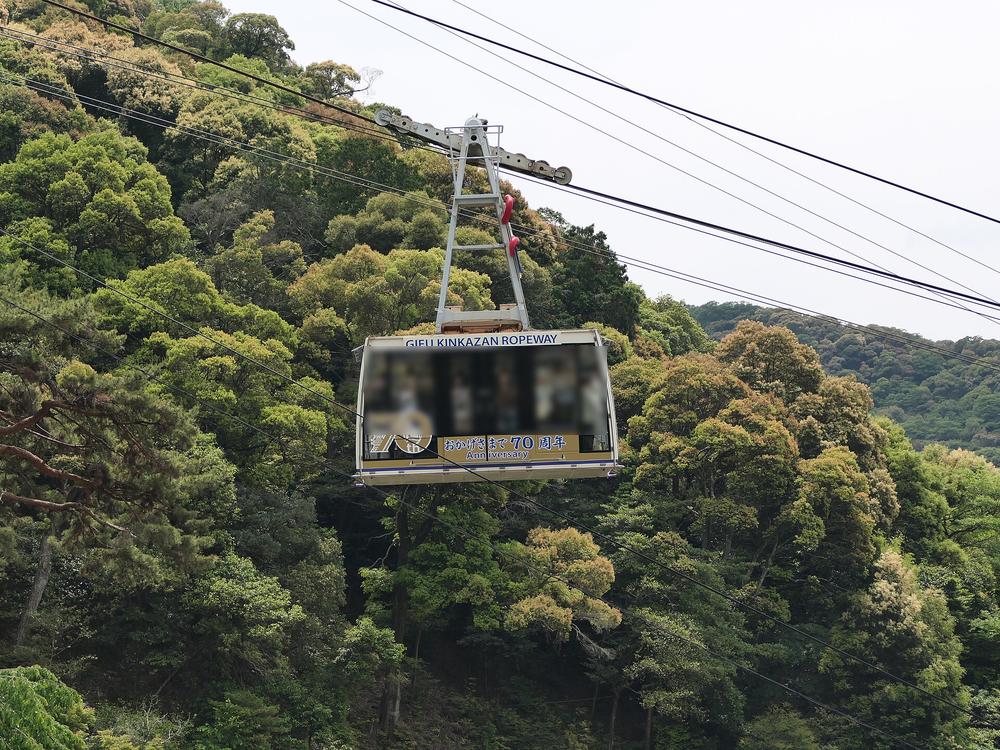 金華山纜車車廂懸在纜索上,背景是整片綠色山林;車廂上方寫著 GIFU KINKAZAN ROPEWAY,下方是「おかげさまで 70 周年」的標示