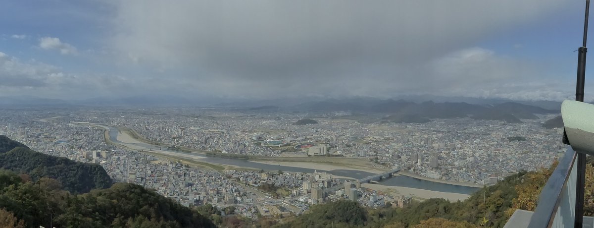 從金華山展望台往北看的全景:長良川從左往右彎過岐阜市區,河上有兩座橋,河岸是大片白色礫石灘,遠方是一排山脈,右緣看得到展望台的欄杆和投幣望遠鏡