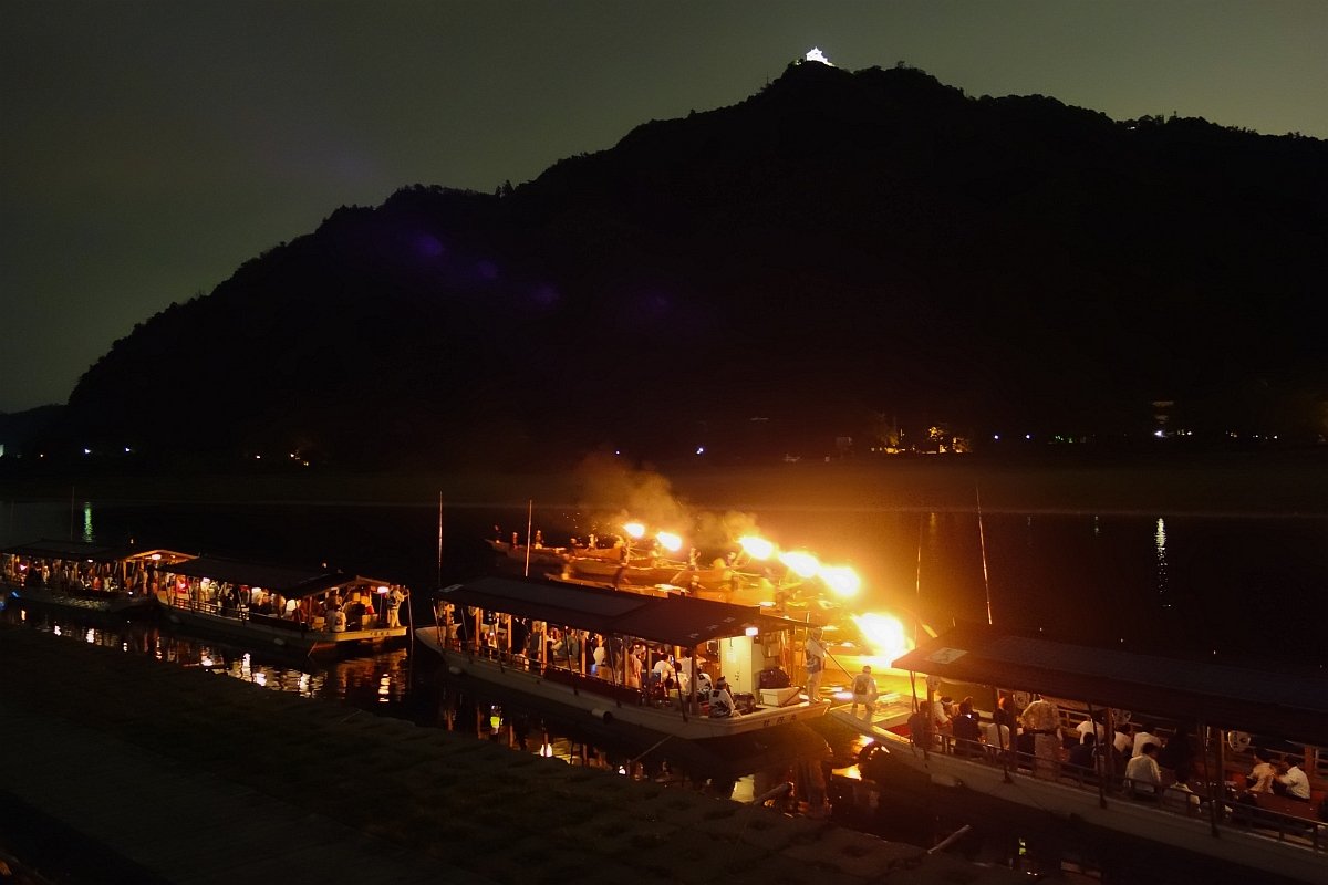 夜晚的長良川鵜飼:河面上幾艘鵜舟的篝火燒得通紅、冒著煙,旁邊停靠著坐滿遊客的屋形觀覽船,背後是漆黑的金華山,山頂有一點打了燈的天守