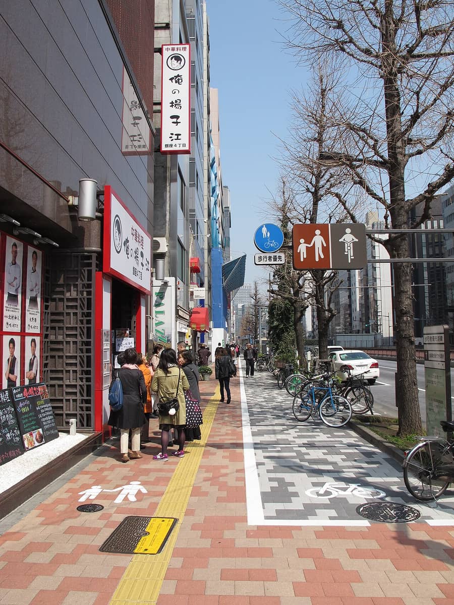 東京銀座四丁目的人行道:桿上一面藍底圓形標識畫著大人牽小孩與一台單車,下方白色補助牌逐字「歩行者優先」,旁邊一面咖啡色雙格圖示左邊是行人、右邊是騎士;地面被白線切成兩半,左半紅磚色鋪面畫白色行人圖示、有一排人在店門口排隊,右半灰色花紋鋪面畫白色單車圖示並緊鄰車道