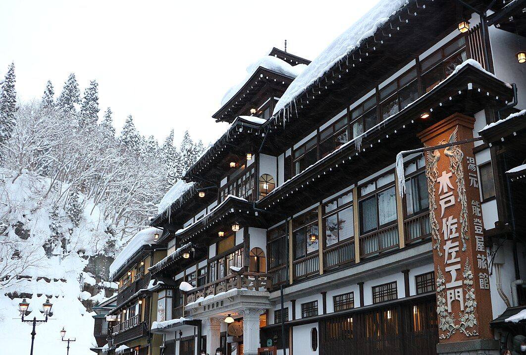 雪中的能登屋旅館：三層木造建築的屋簷壓著厚雪、垂著冰柱，走廊一排吊燈亮著，右側立著刻有鳳凰浮雕的「木戸佐左エ門」看板與「湯元 能登屋旅館」直式招牌