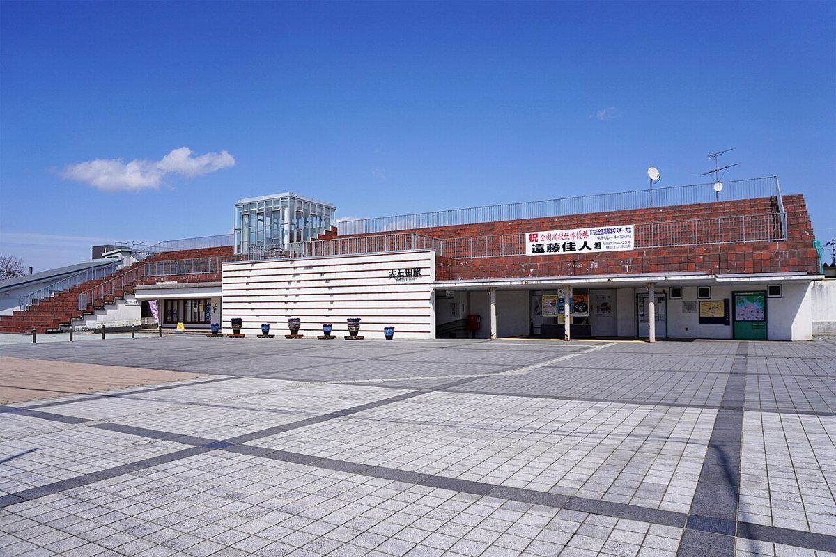 JR 大石田站西口站舍：紅磚色斜屋頂搭白色橫條牆面，牆上寫著「大石田駅」，站前是一大片空曠的方磚廣場
