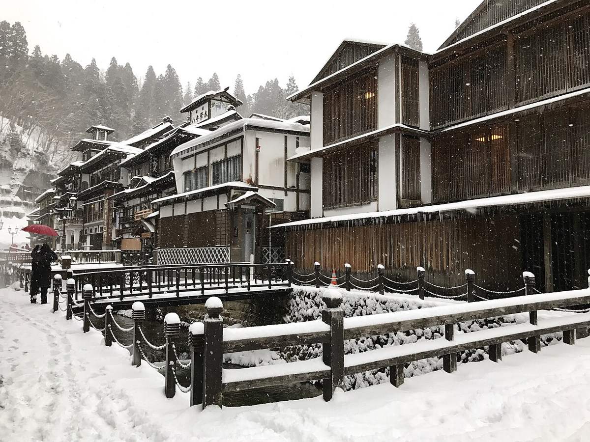 下雪中的銀山溫泉街步道：地面是被踩實的雪、留著一道道腳印，河岸鐵鍊欄杆與對岸旅館的木格柵全被雪蓋住，左後方一個撐紅傘的人背對鏡頭走著