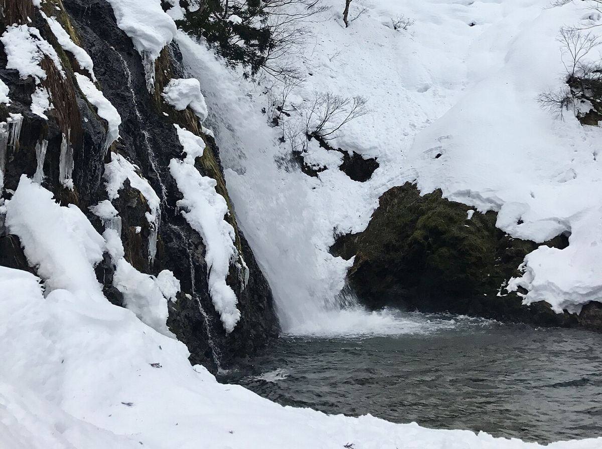 冬天的白銀之瀧：水流從左側黑色岩壁落進深色水潭，岩壁掛著冰柱、岩頂與右側斜坡覆蓋厚雪，前景的雪地滿是腳印