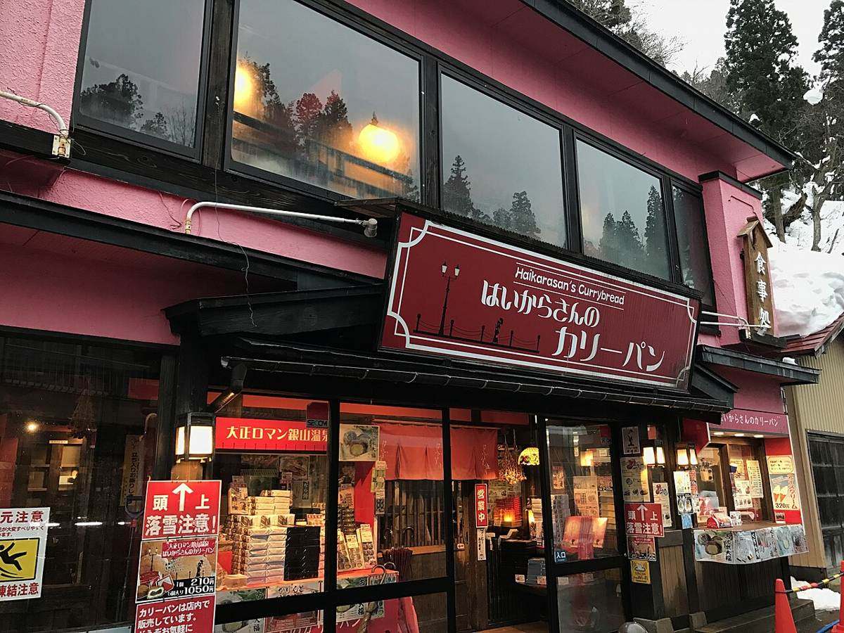 銀山溫泉街上一間粉紅色外牆的店面，門口立著紅底白字的「頭上 落雪注意！」告示牌，左邊另有黃底的「凍結注意」滑倒圖示牌，屋簷與後方山坡都積著雪