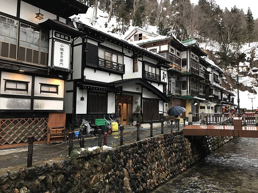 銀山溫泉街旁的木造旅館「小関吉左衛門」門口，停著一台送貨機車、兩台手推台車和幾個綠色塑膠箱，右前方是紅欄杆小橋與銀山川