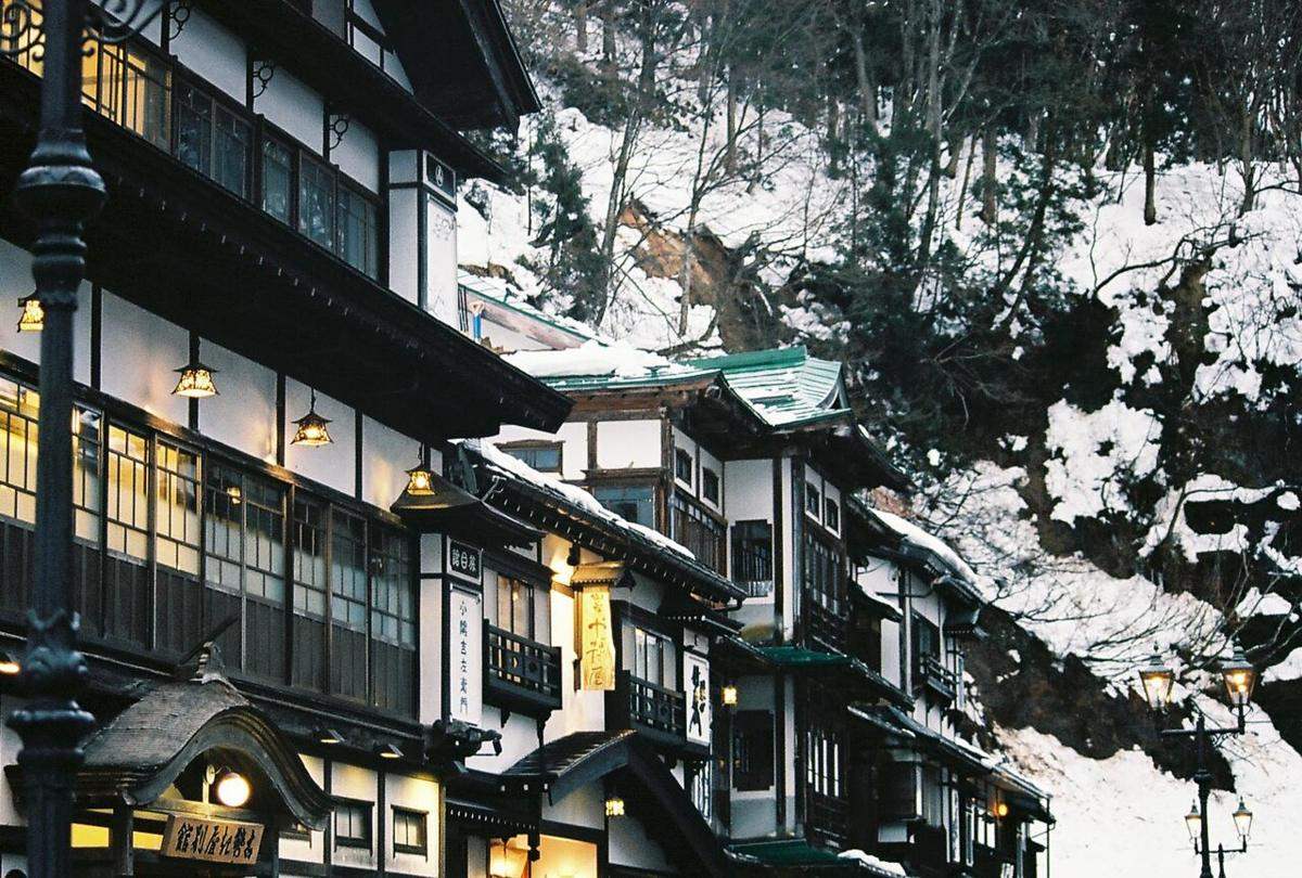 天色轉暗的銀山溫泉街：木造旅館二樓走廊的吊燈與房間窗戶亮著暖黃光，畫面左緣和右側各有一盞造型煤氣燈已點亮，背後山坡全是積雪