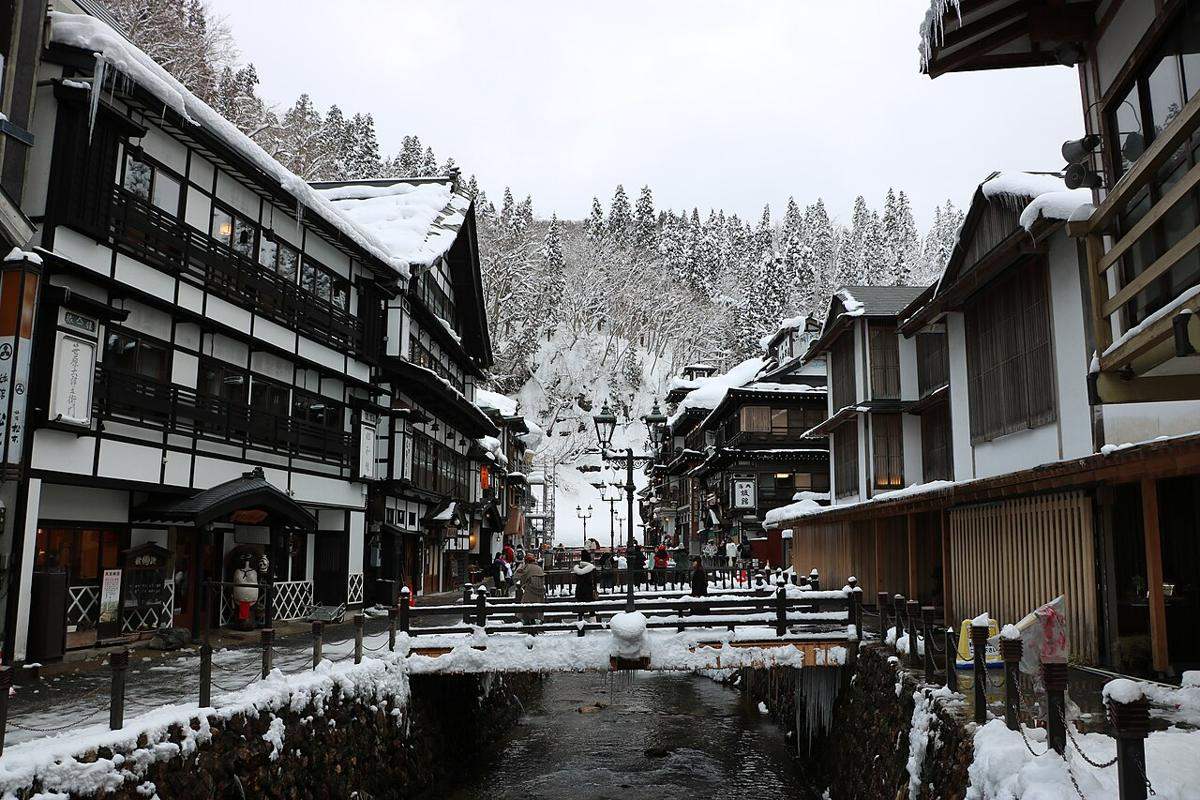 下雪後的銀山溫泉街：銀山川兩側木造旅館屋簷厚厚積雪，兩岸各只有一條窄窄的行人道，遠處小橋上有幾個遊客，路面上沒有任何車輛