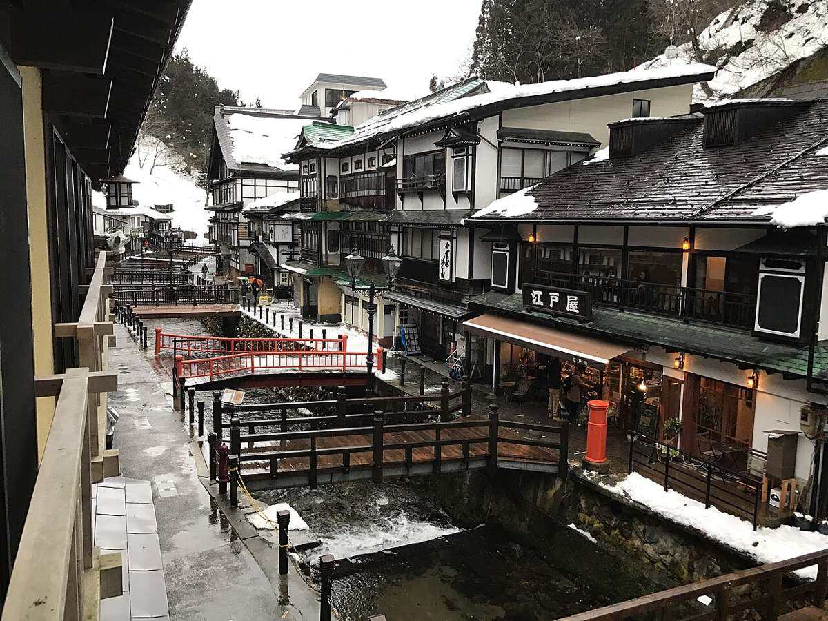 從旅館二樓外廊俯看銀山溫泉街：銀山川夾在兩排木造旅館中間，河上有一座紅欄杆小橋與兩座木橋，右側「江戸屋」招牌下是一排店面，屋頂與河岸殘雪未化