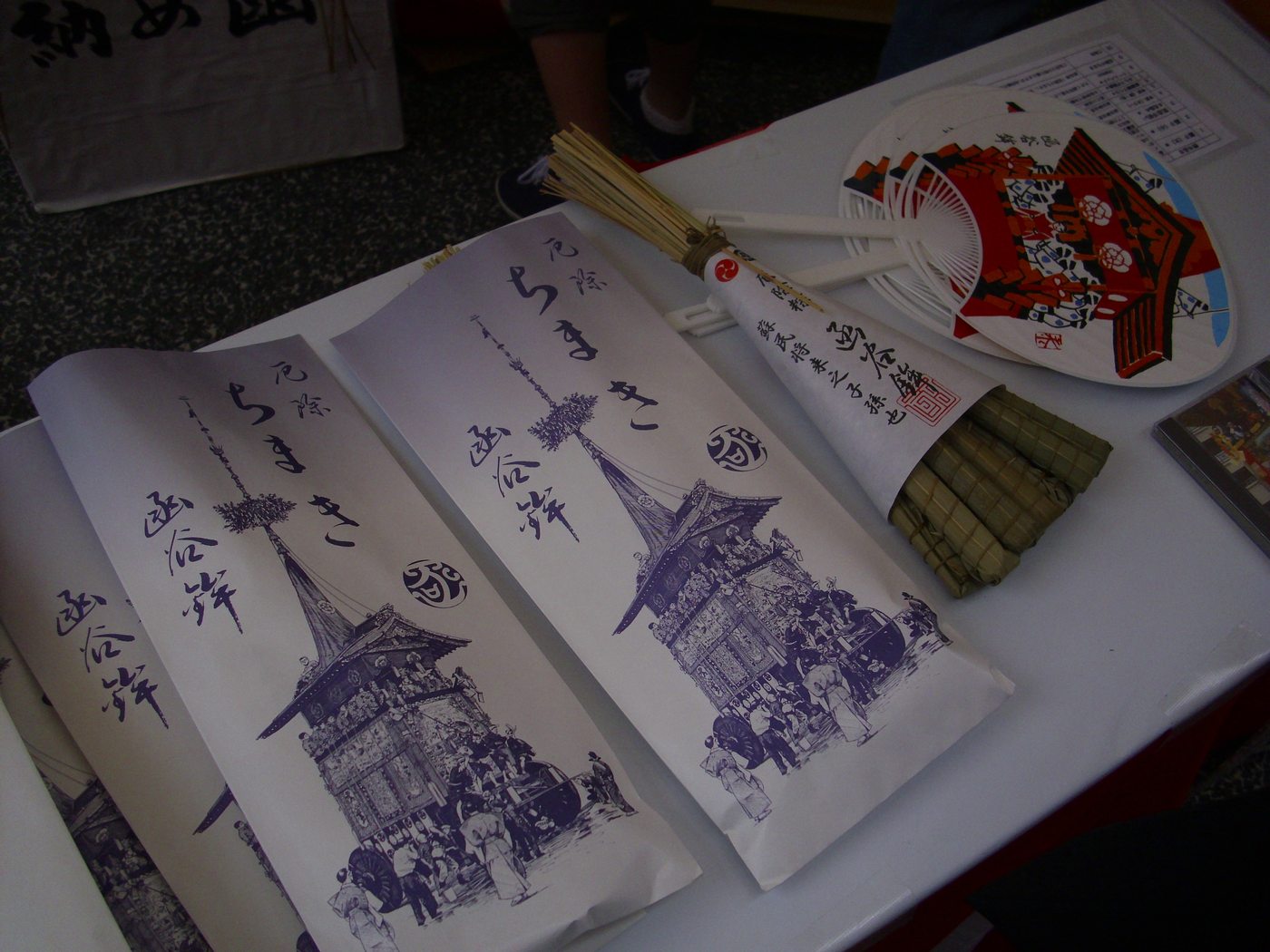 鋪著白布的桌上攤著幾個印有「厄除ちまき 函谷鉾」的長條紙袋,右邊露出一束以笹葉綑成的粽,旁邊放著紙團扇